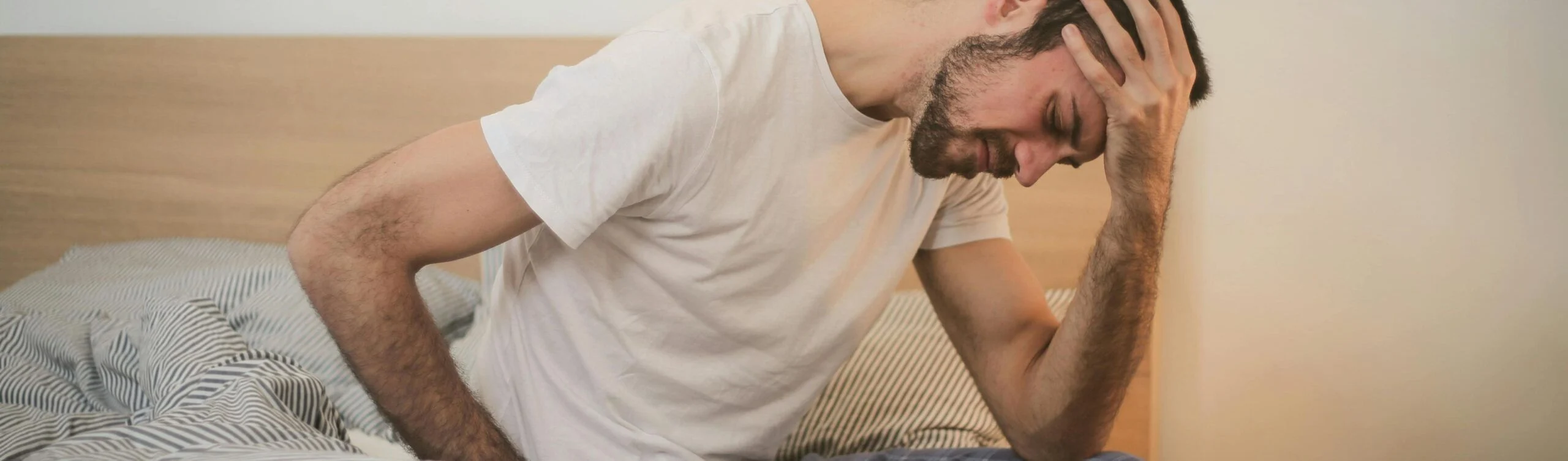 Una fotografía de plano medio que muestra a un hombre joven sentado en el borde de una cama desordenada en una habitación con iluminación tenue. Viste un pijama compuesto por una camiseta blanca de manga corta y pantalones de cuadros azules. El hombre aparece encorvado, con su mano derecha presionando su frente en un gesto de intenso dolor, frustración o desesperación, mientras su mano izquierda descansa sobre su espalda baja. La expresión de su rostro, parcialmente oculta, refleja agotamiento físico y mental. Al fondo se aprecia el cabecero de madera de la cama y una mesita de noche blanca, creando una atmósfera de soledad y reflexión íntima.