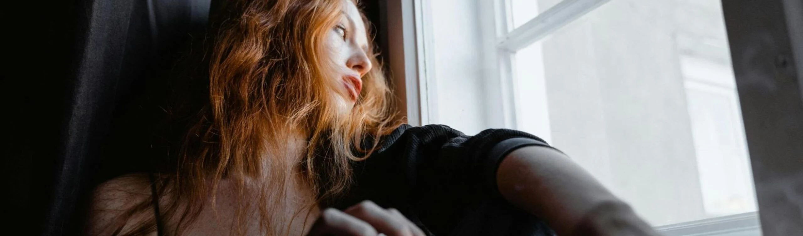 Una fotografía de plano medio capturada desde un ángulo bajo que muestra a una mujer joven de cabello pelirrojo largo y ondulado. Se encuentra en una habitación con poca luz, situada junto a una ventana por la que entra una claridad fría y difusa. La mujer viste una prenda negra y mira hacia el exterior con una expresión de profunda melancolía, ausencia y cansancio. En primer plano, su mano derecha aparece ligeramente desenfocada cerca del marco de la ventana, subrayando la sensación de encierro y la desconexión con el mundo real que caracteriza a las adicciones digitales tratadas en Bella Mar.