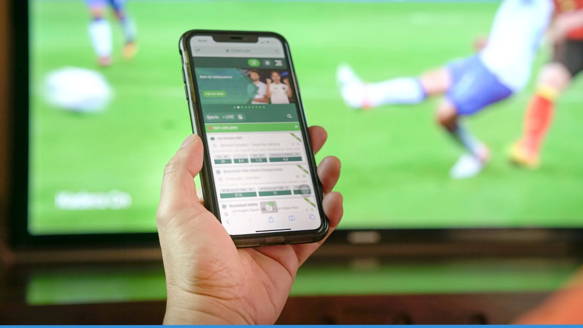 Una fotografía de primer plano que muestra la mano de una persona sosteniendo un teléfono móvil inteligente (smartphone) frente a una pantalla de televisión de grandes dimensiones. En la pantalla del teléfono se visualiza una aplicación de apuestas deportivas con listas de cuotas y mercados de eventos en vivo. Al fondo, la televisión muestra un partido de fútbol desenfocado donde se ve a un jugador en acción sobre el césped verde. La imagen captura la conexión inmediata entre el consumo de contenido deportivo y la facilidad de acceso al juego digital, ideal para ilustrar los disparadores de la ludopatía en Bella Mar.