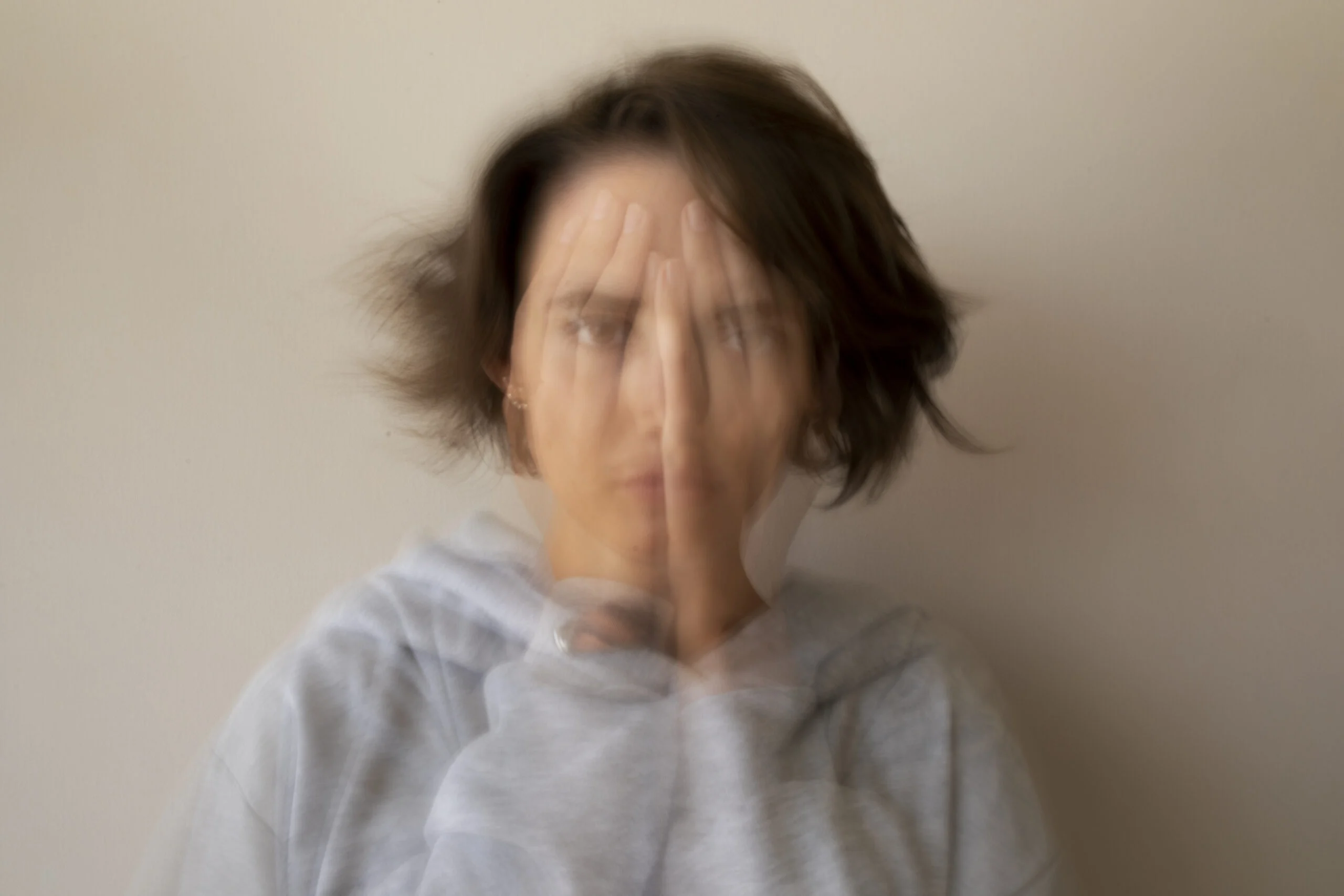 Retrato de plano medio con un efecto de larga exposición o doble exposición que crea un desenfoque de movimiento. En la imagen, una persona aparece con las manos frente al rostro, pero la distorsión visual hace que las facciones y las manos se multipliquen en capas semitransparentes. Proyectando una sensación de inestabilidad, agitación interna y la dificultad de mantener el enfoque en medio de una crisis de ansiedad.